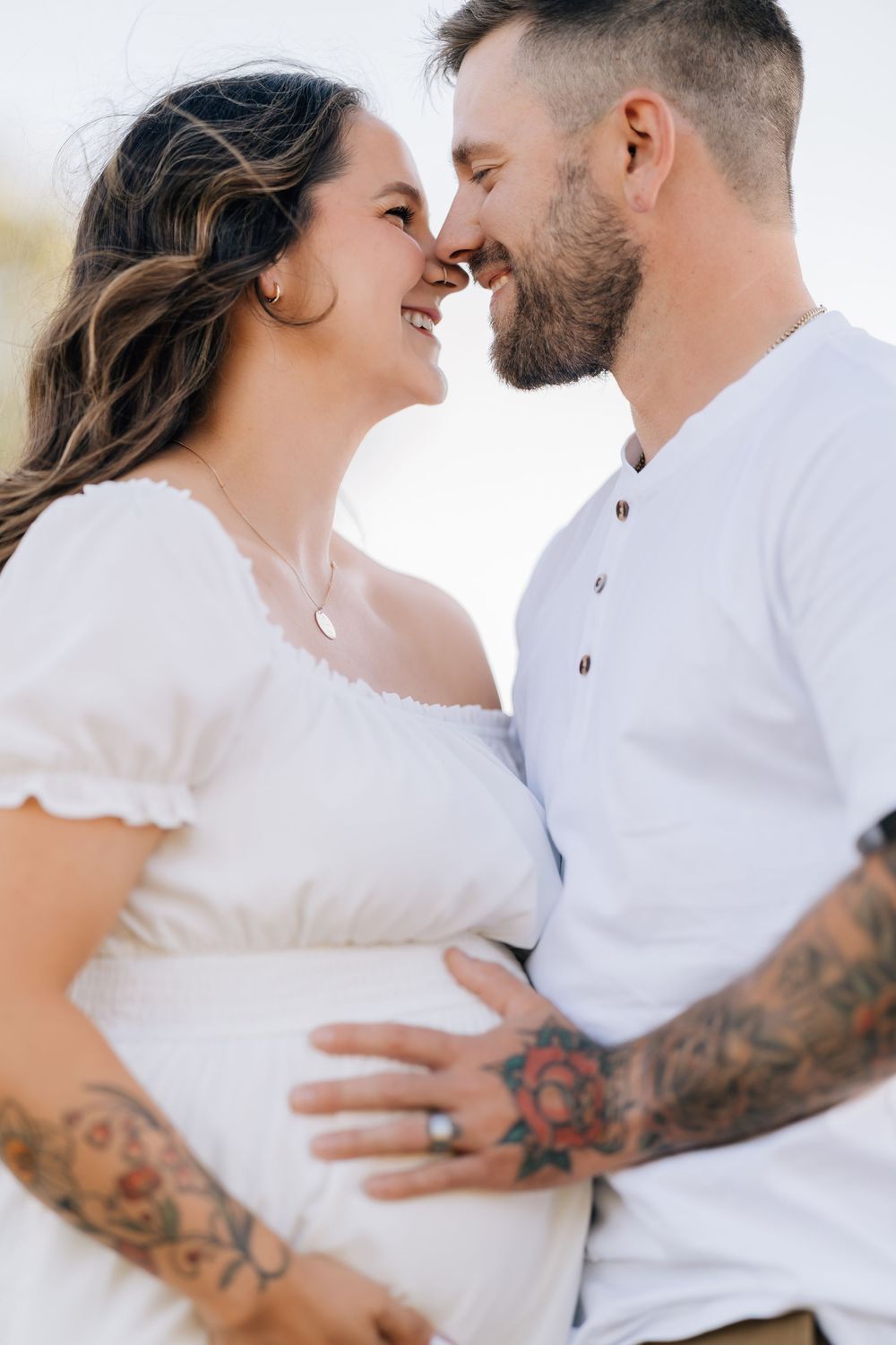 Smiling pregnant couple in white outfits touching noses romantically, man with tattoos cradling baby bump outdoors.
