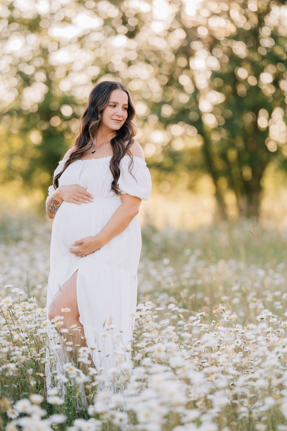 Pregnant woman in white dress standing in daisy field at golden hour, cradling baby bump with gentle smile.