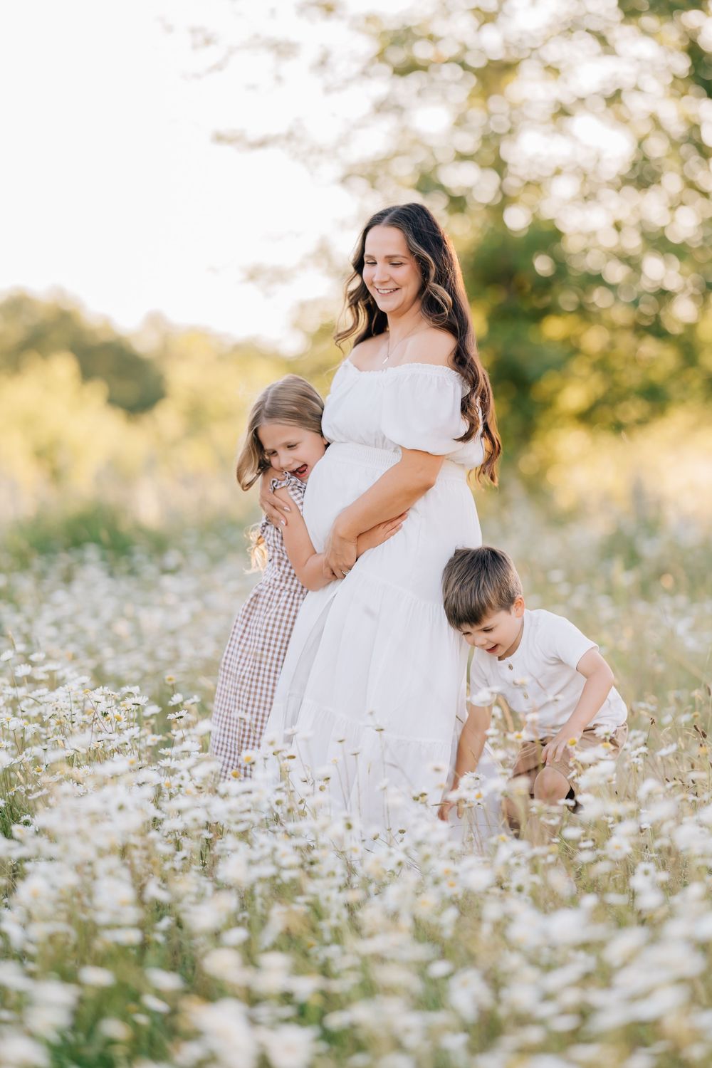Pregnant woman in white off-shoulder dress standing in daisy field with two young children hugging her bump.