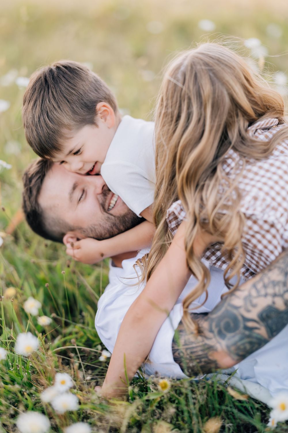 Tattooed father laughing joyfully as young son kisses his cheek while lying in a sunny wildflower meadow.