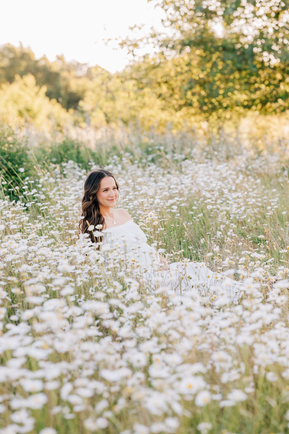 A smiling woman in a white dress sits surrounded by blooming white daisies in a sunlit meadow.