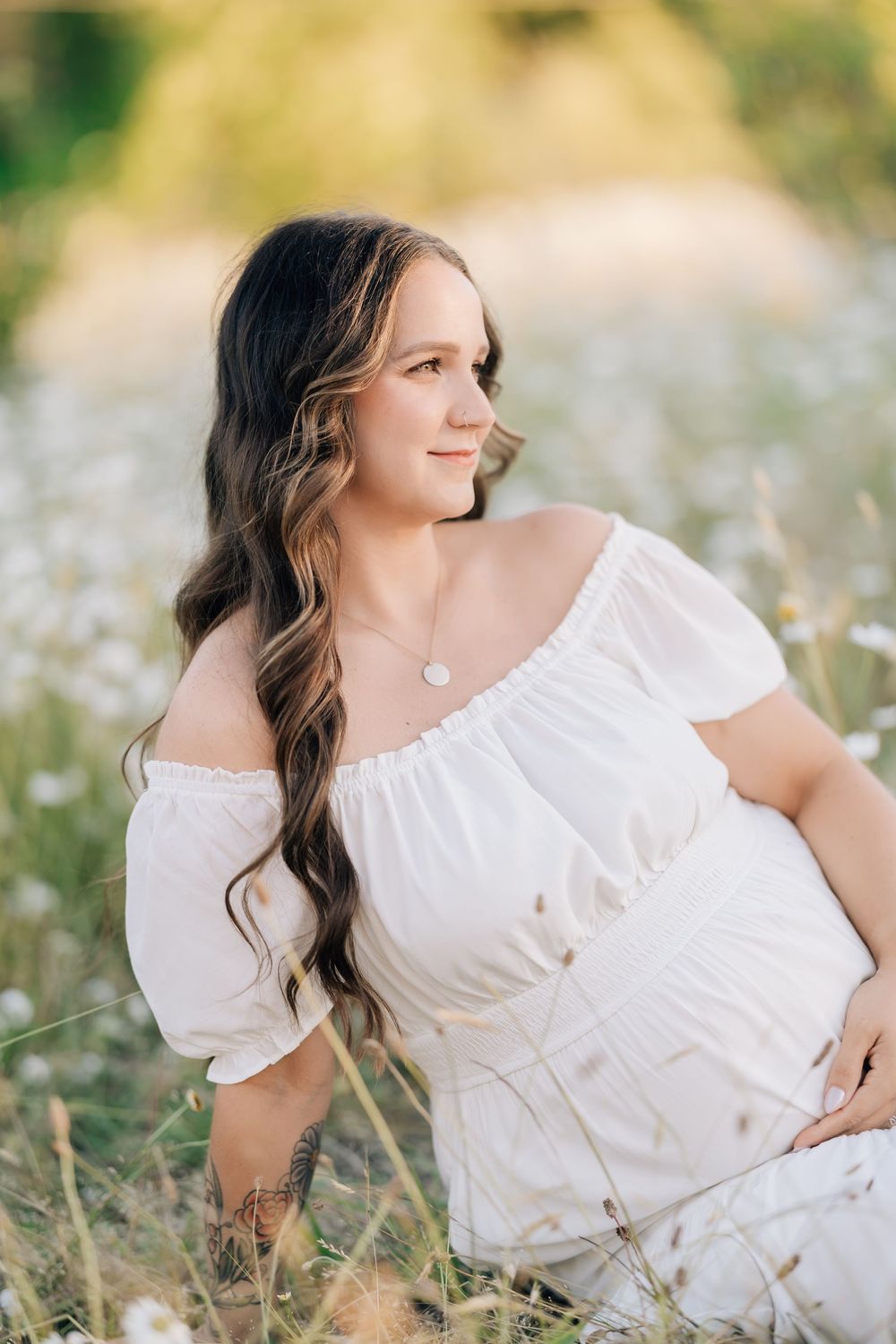 Beautiful pregnant woman in off-shoulder white dress sitting in wildflower field during golden hour sunset.