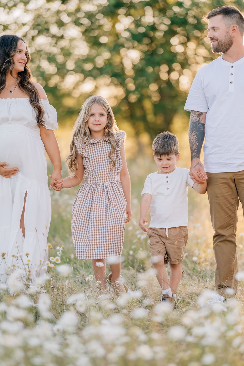 A happy pregnant family of four walks hand in hand through a sunny wildflower field.