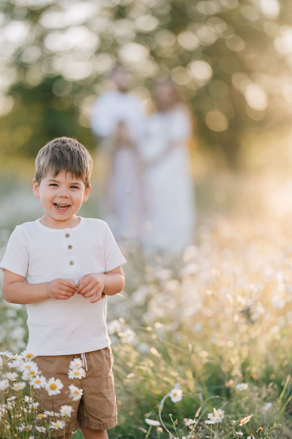 Happy toddler boy smiling in daisy field with parents in background at sunset.
