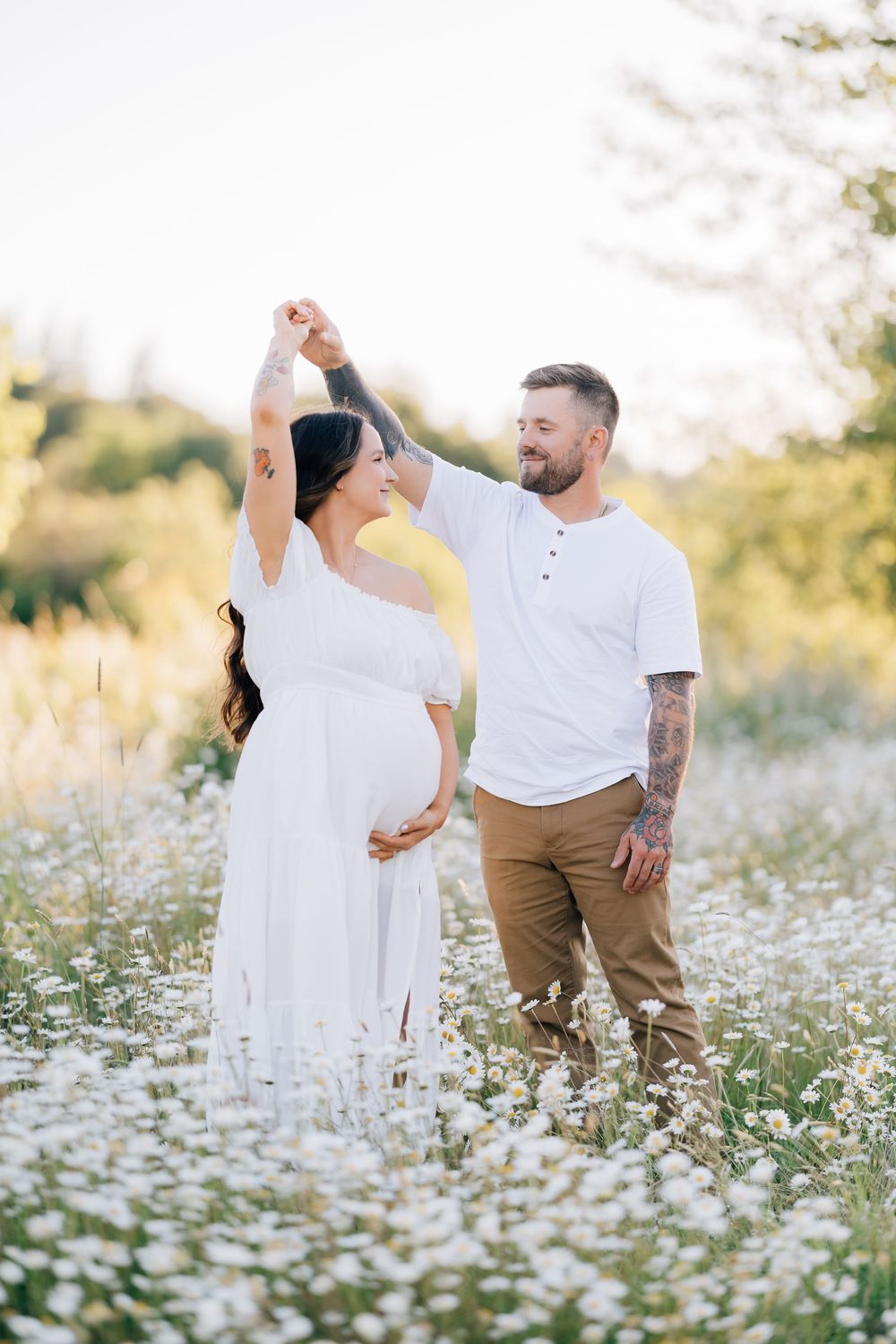 Pregnant woman in white dress dances with tattooed man in daisy field during golden hour maternity photoshoot.
