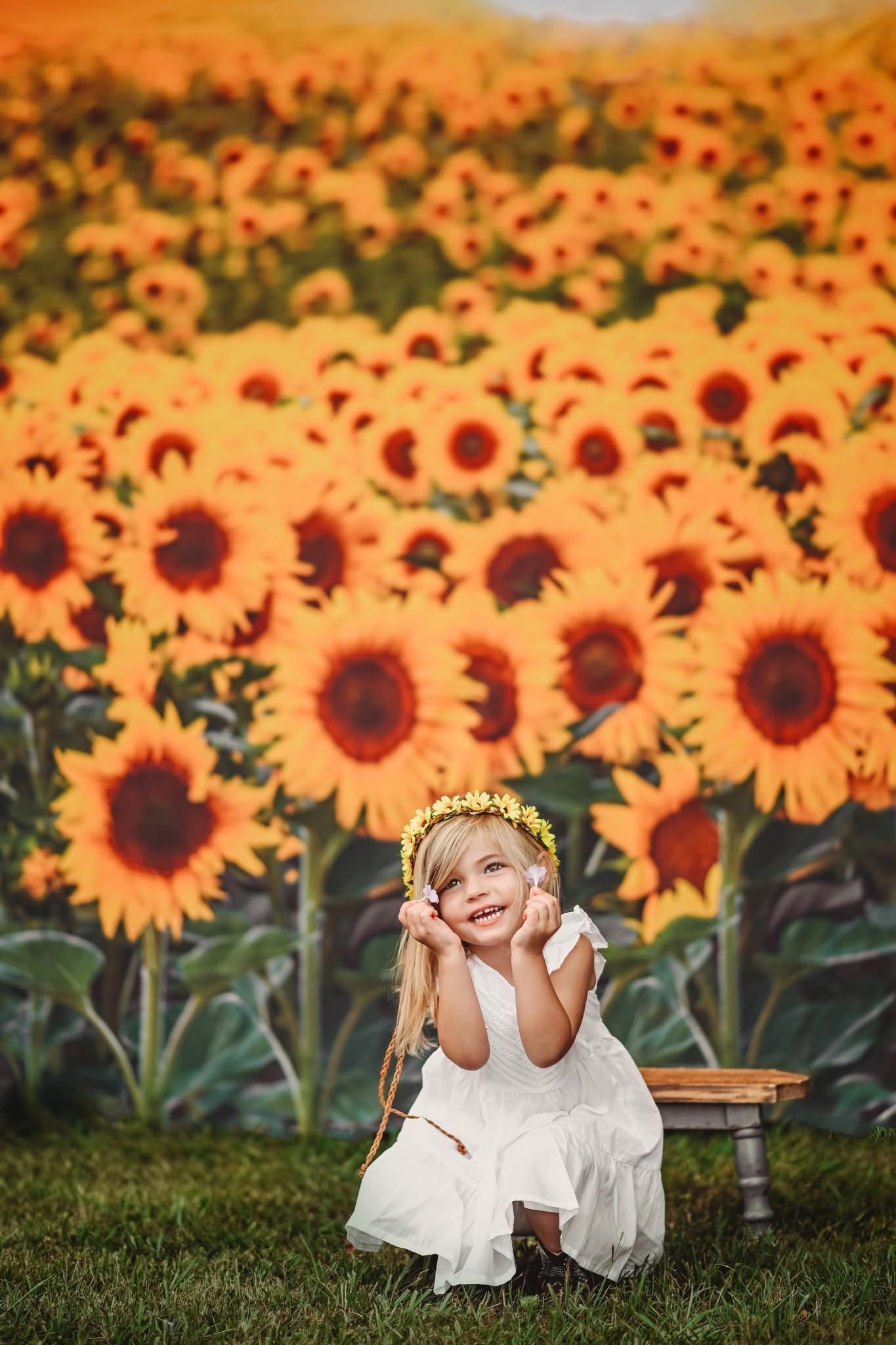 Young girl in white dress and floral crown sits smiling before a vibrant sunflower field backdrop.