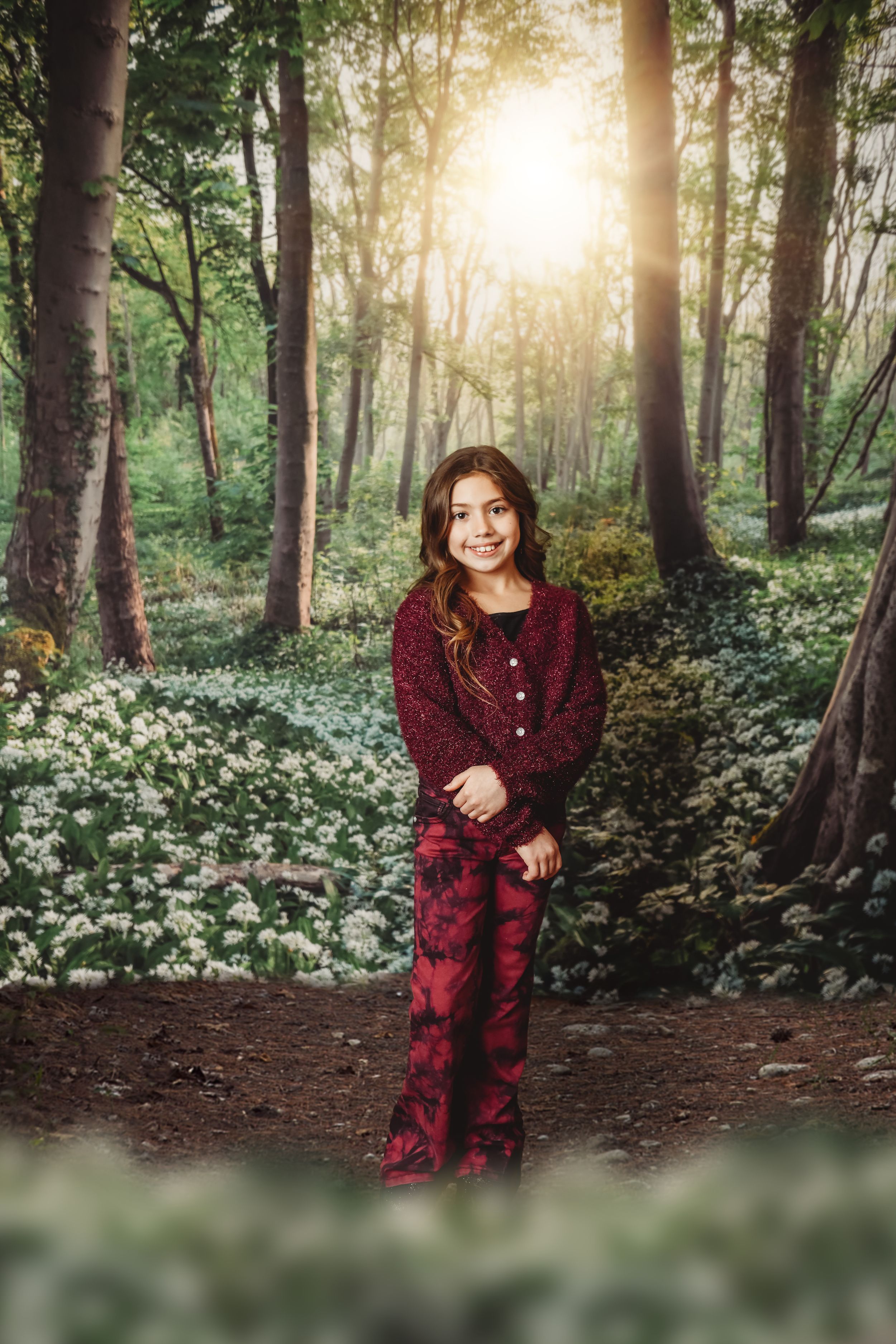 A smiling girl in a red outfit stands on a forest path surrounded by white wildflowers and sunlit trees.