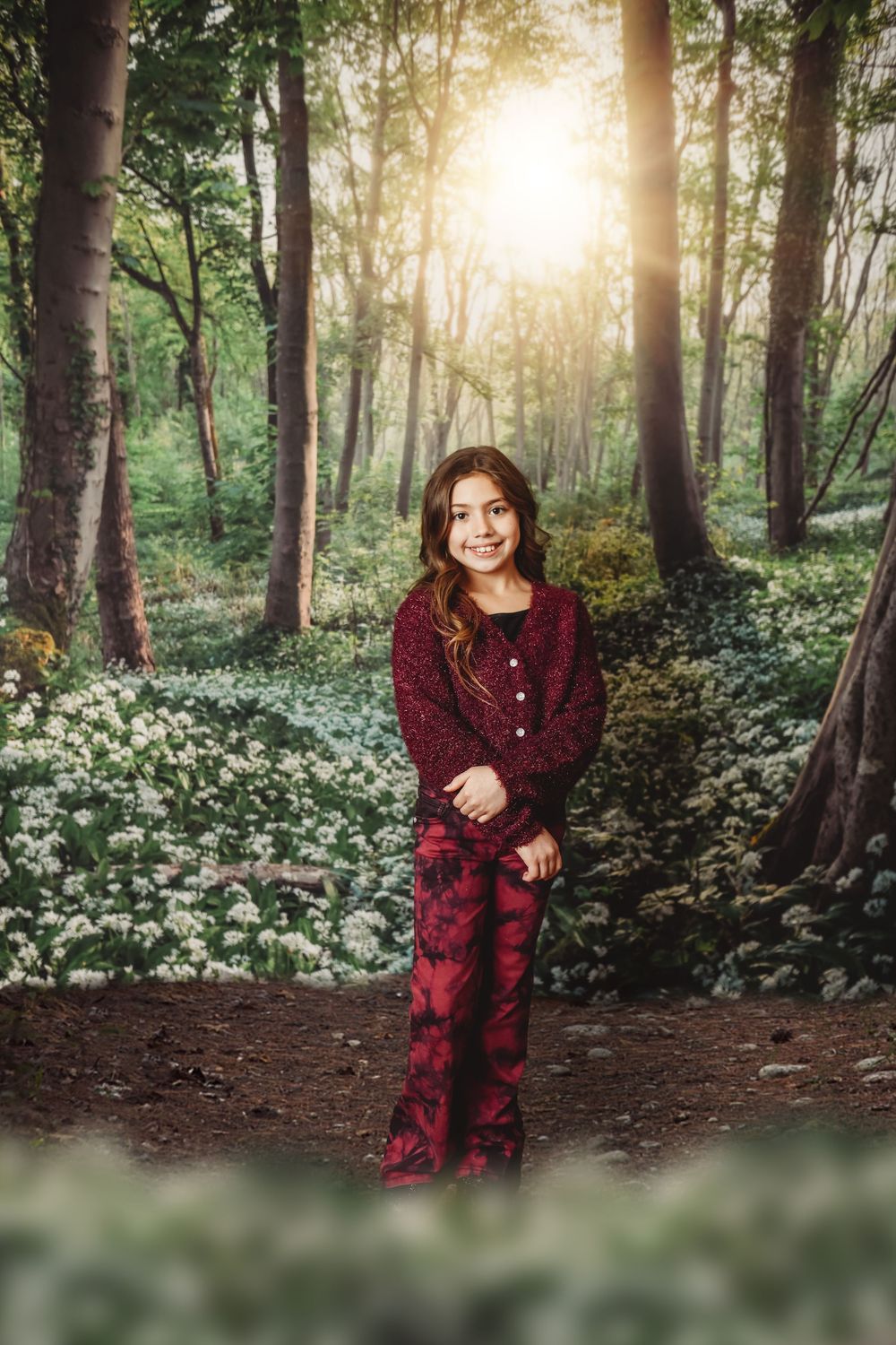 A smiling girl in a red outfit stands on a forest path surrounded by white wildflowers and sunlit trees.