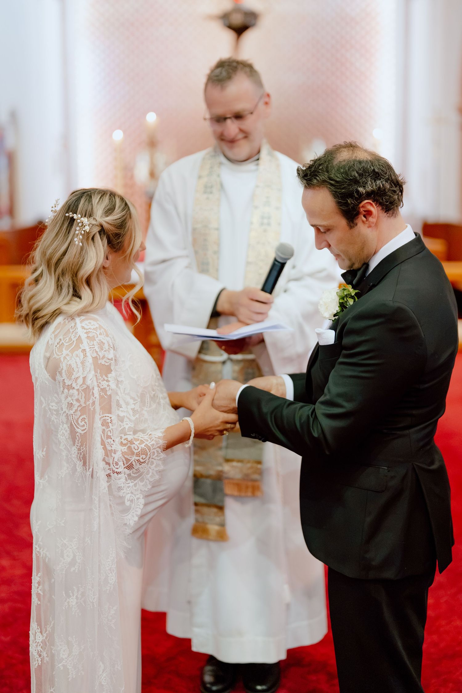 Bride in a lace gown and groom in a black suit hold hands during their wedding ceremony as an officiant reads from a book.