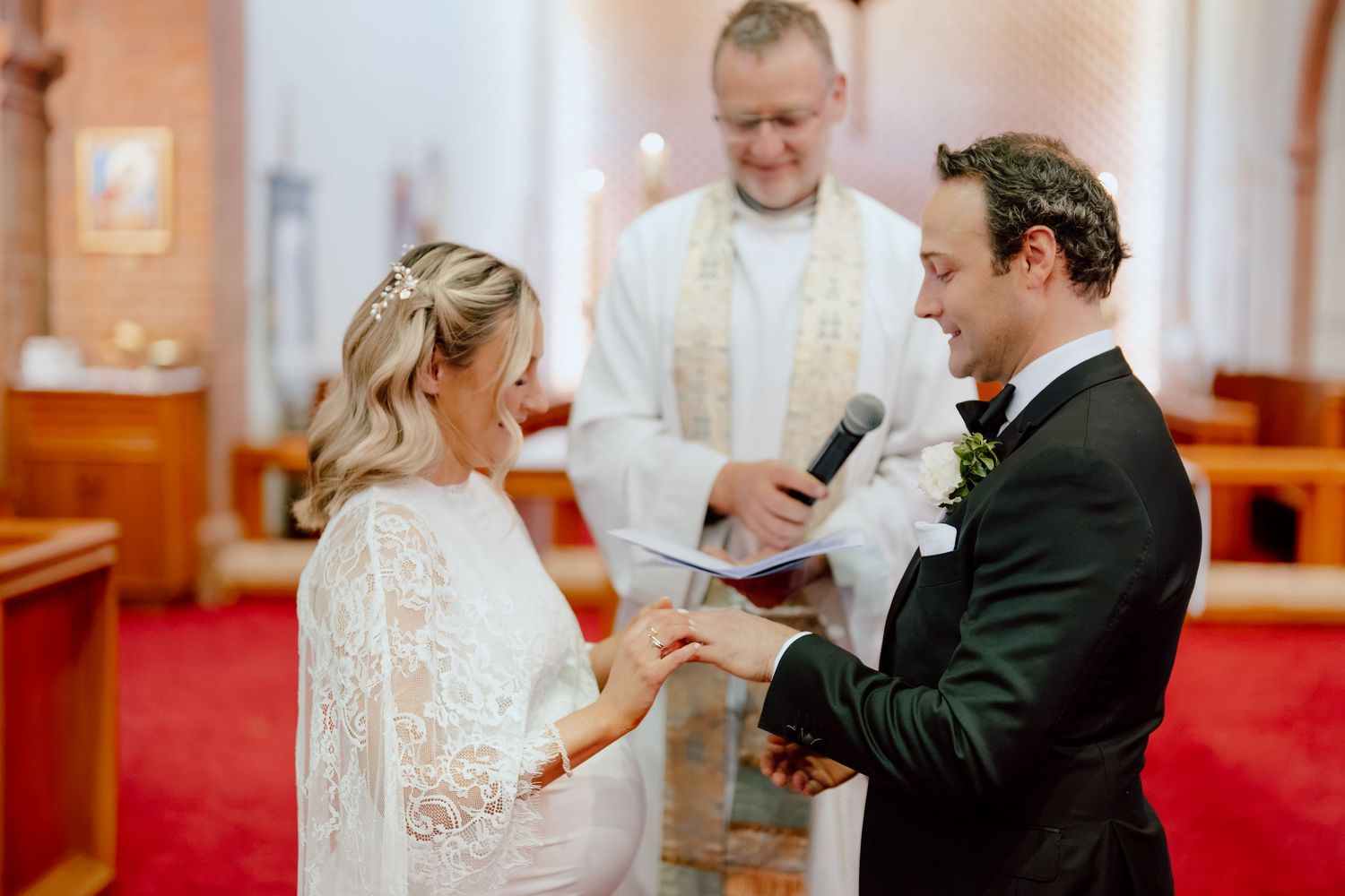 Bride in a white lace dress and groom in a dark suit exchange vows and rings during a church wedding ceremony while an officiant reads.