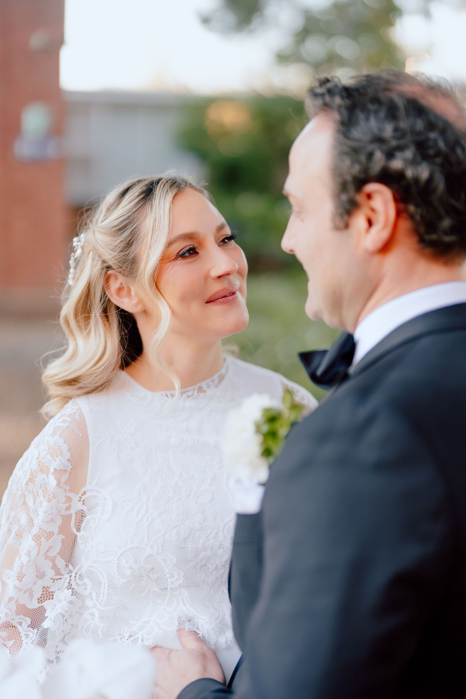 Bride in a white lace dress smiles warmly at a groom in a black tuxedo and bow tie during an outdoor wedding portrait.