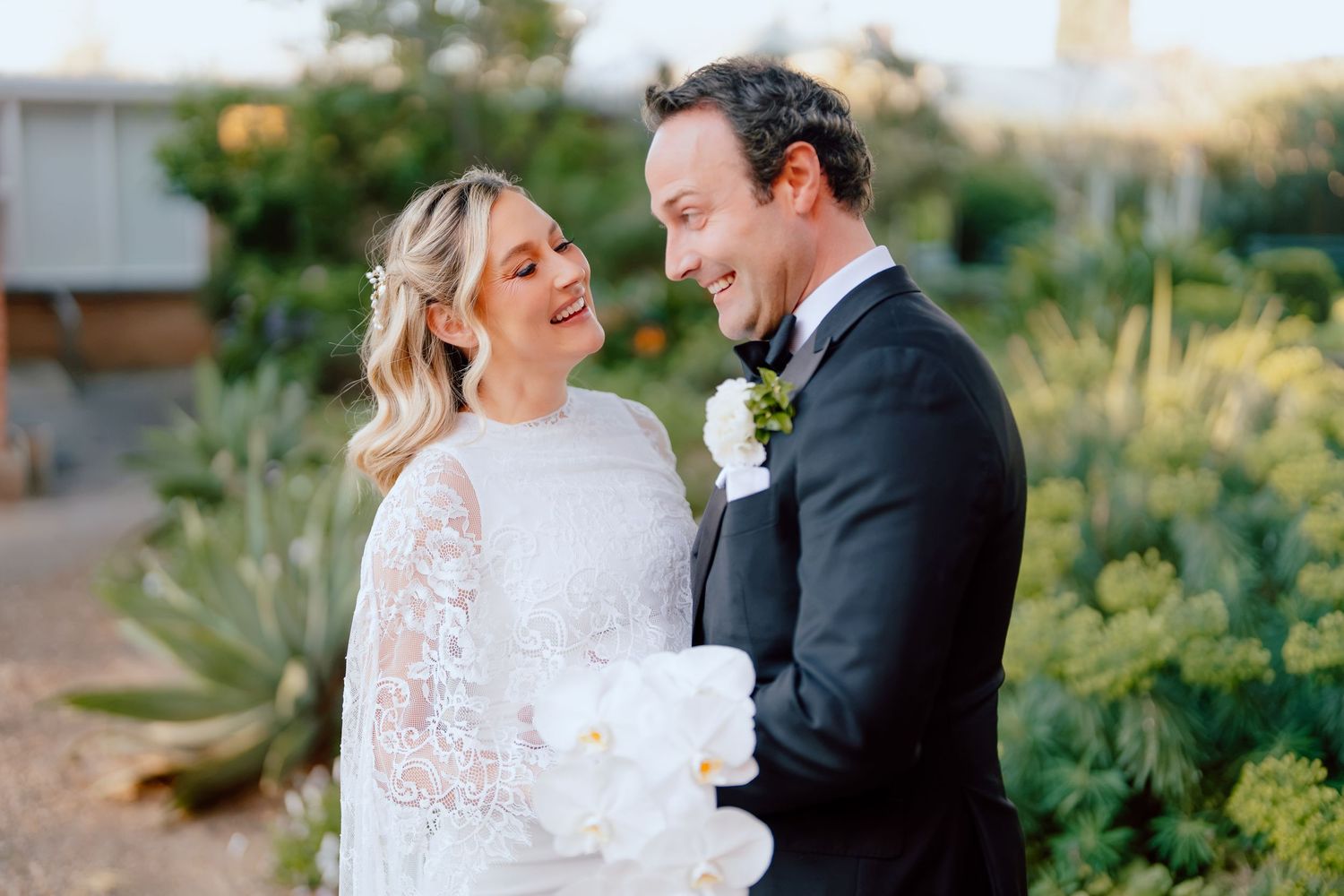 Smiling bride in a white lace wedding dress holding a bouquet embraces her groom in a black tuxedo in a sunny garden.