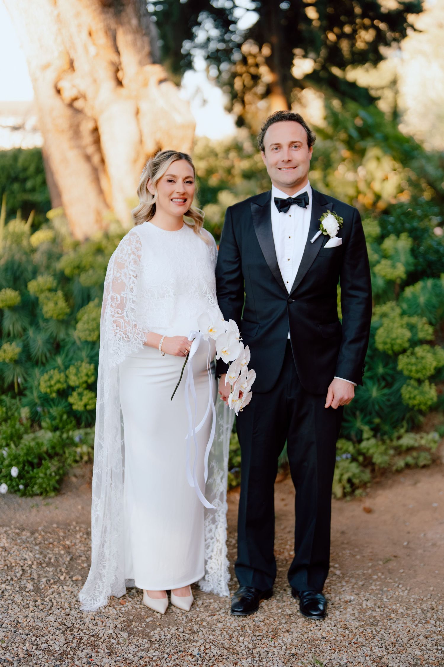 Bride in a white dress with lace cape holding white orchids stands beside a groom in a black tuxedo and bow tie in a garden.