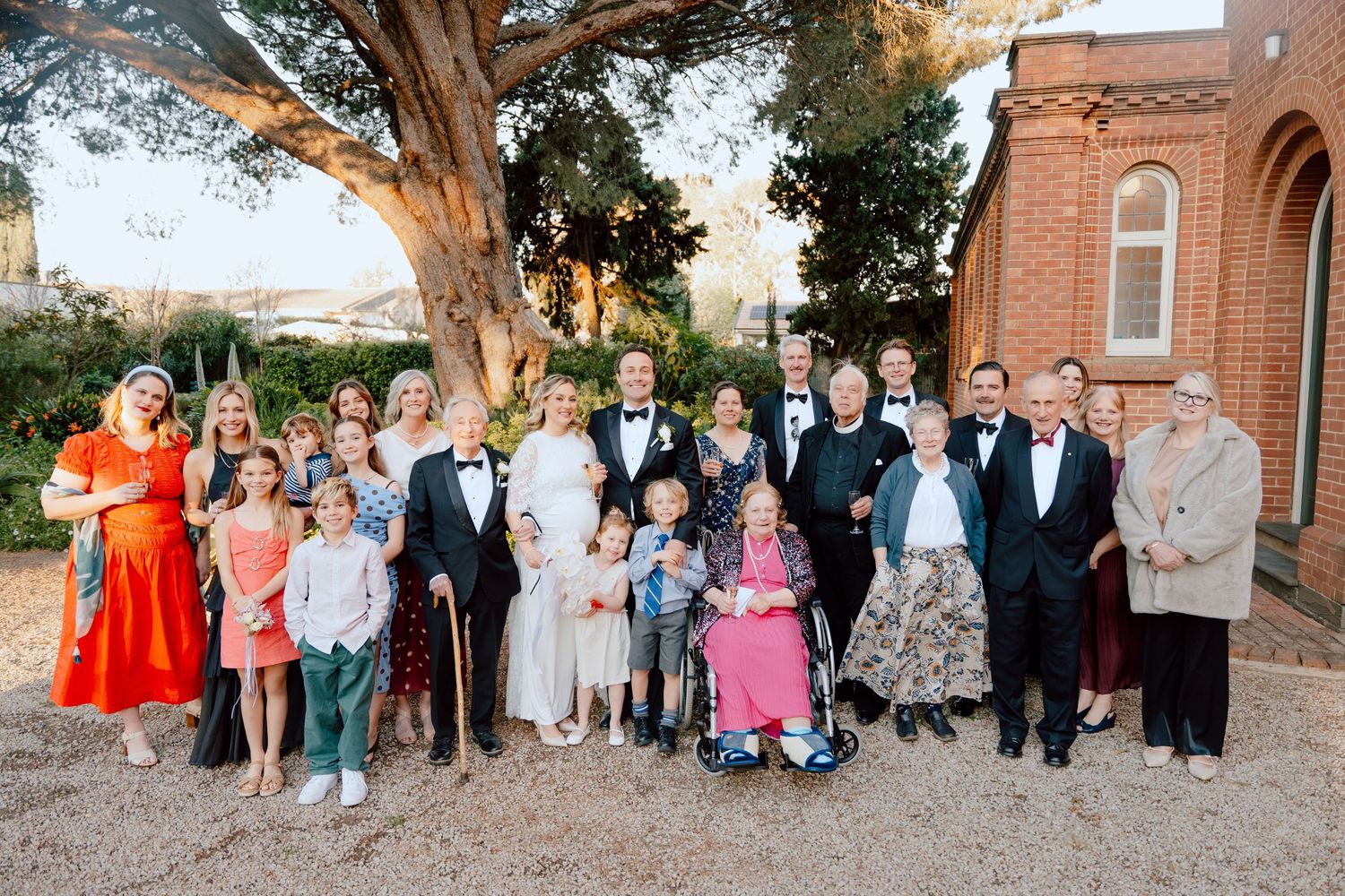Large wedding party group portrait of a bride, groom, and family members in formal attire outside a brick building beneath a tall tree.
