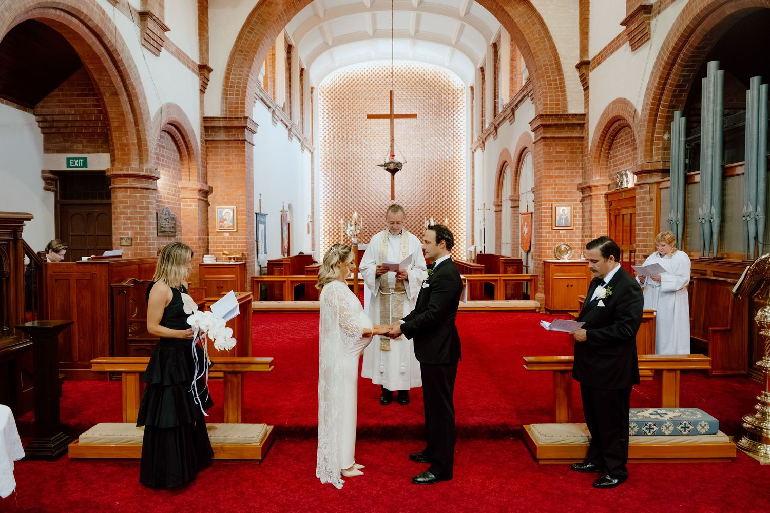 Bride in white dress and groom in black suit exchange vows before an officiant at a red-carpeted church altar with guests looking on.