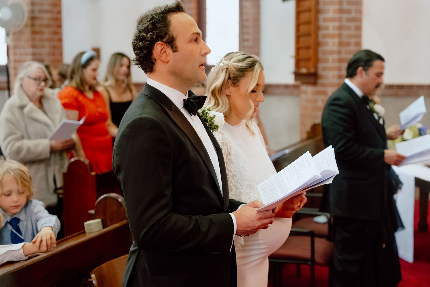 Bride and groom stand side by side holding a program during a church wedding ceremony while guests look on from the pews.