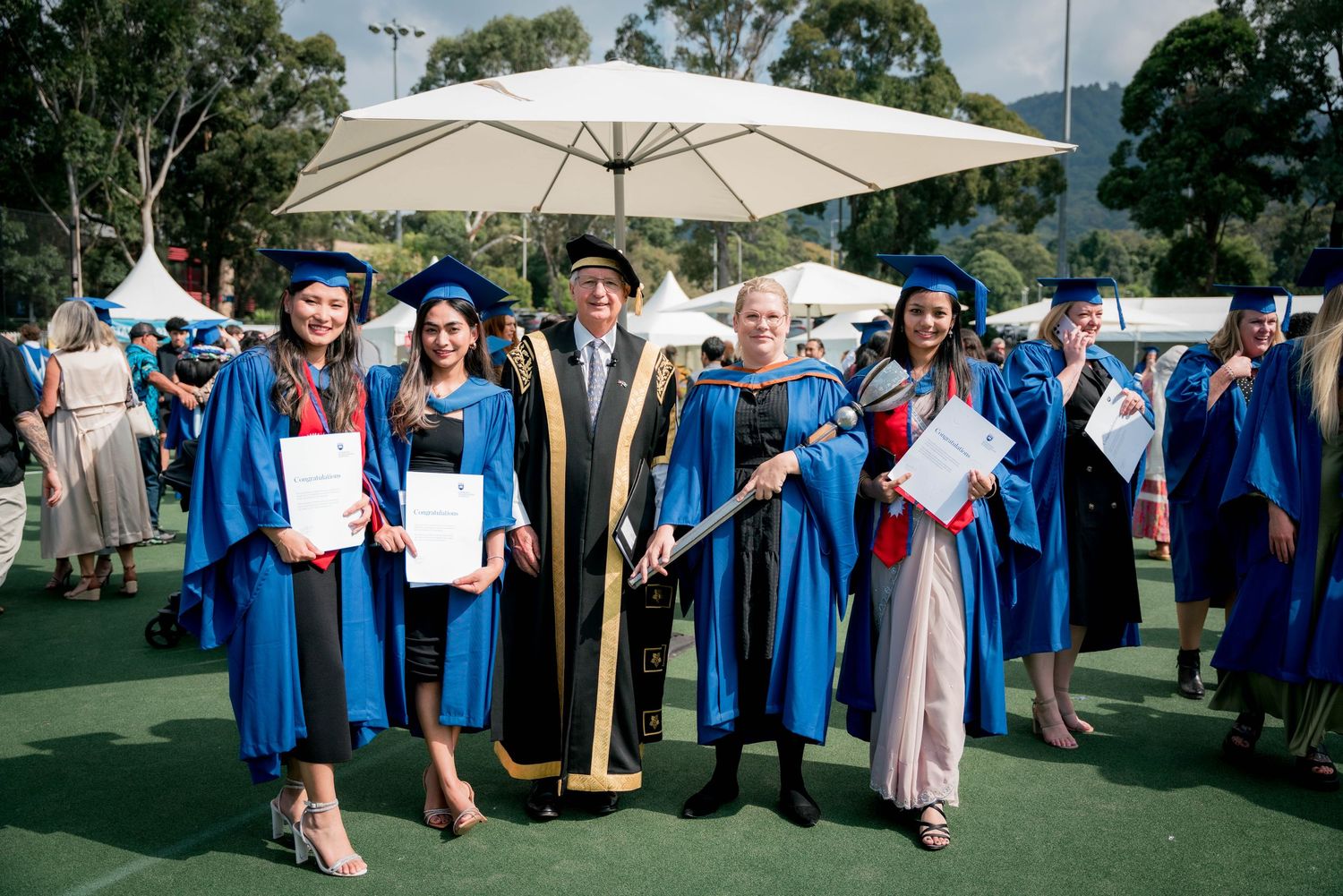 Graduates in blue gowns and caps hold certificates beside an official in a black and gold robe at an outdoor university graduation ceremony.