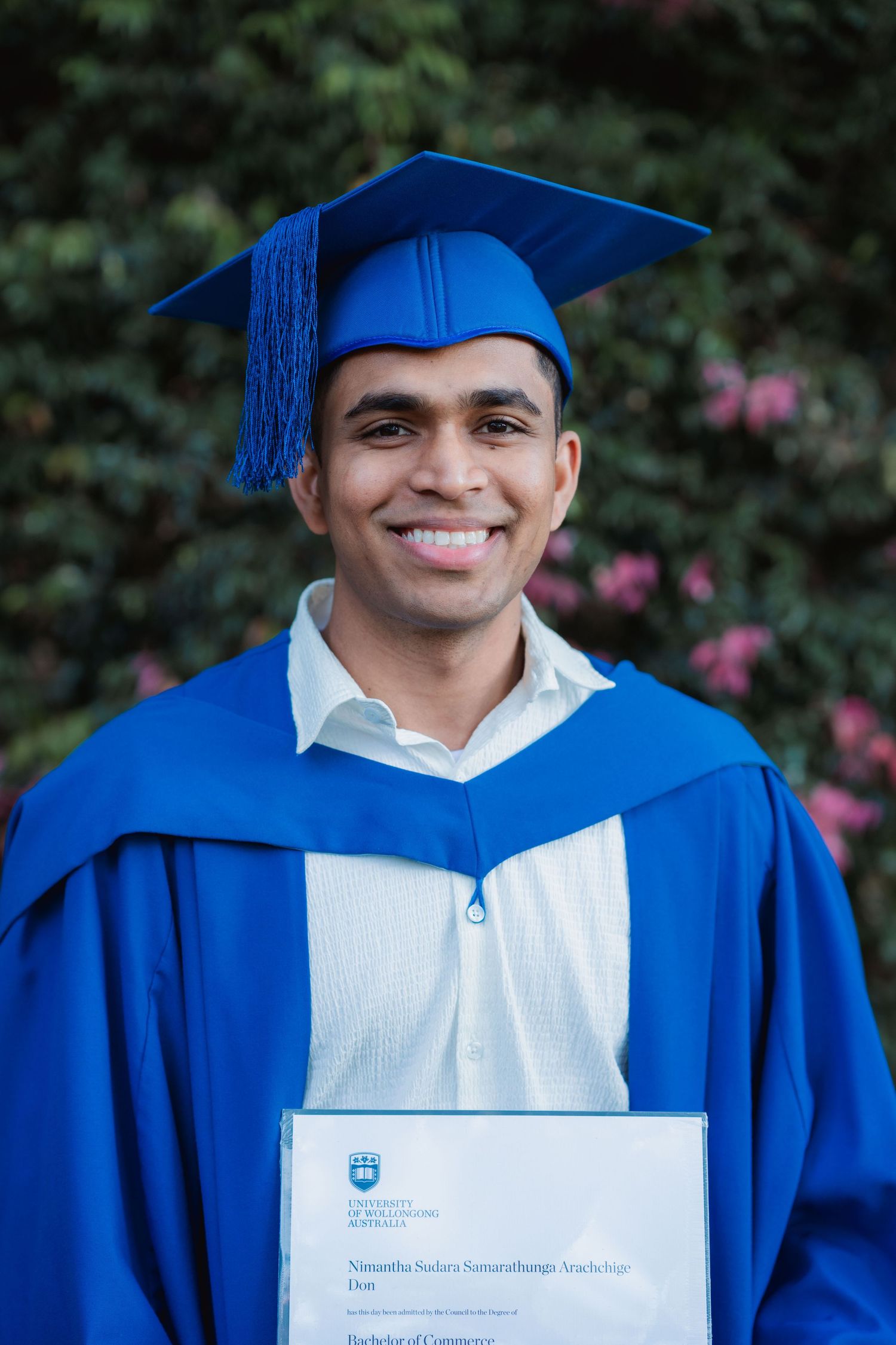 Smiling graduate in a blue cap and gown holds a Bachelor of Commerce certificate in front of green ivy with pink flowers.