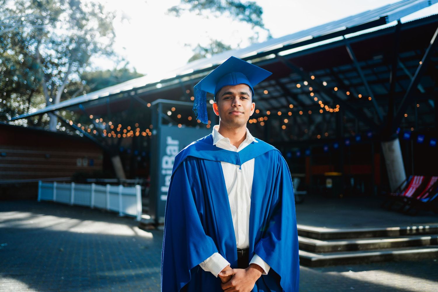 Young graduate in a blue cap and gown stands outdoors with hands clasped, with string lights glowing behind him.