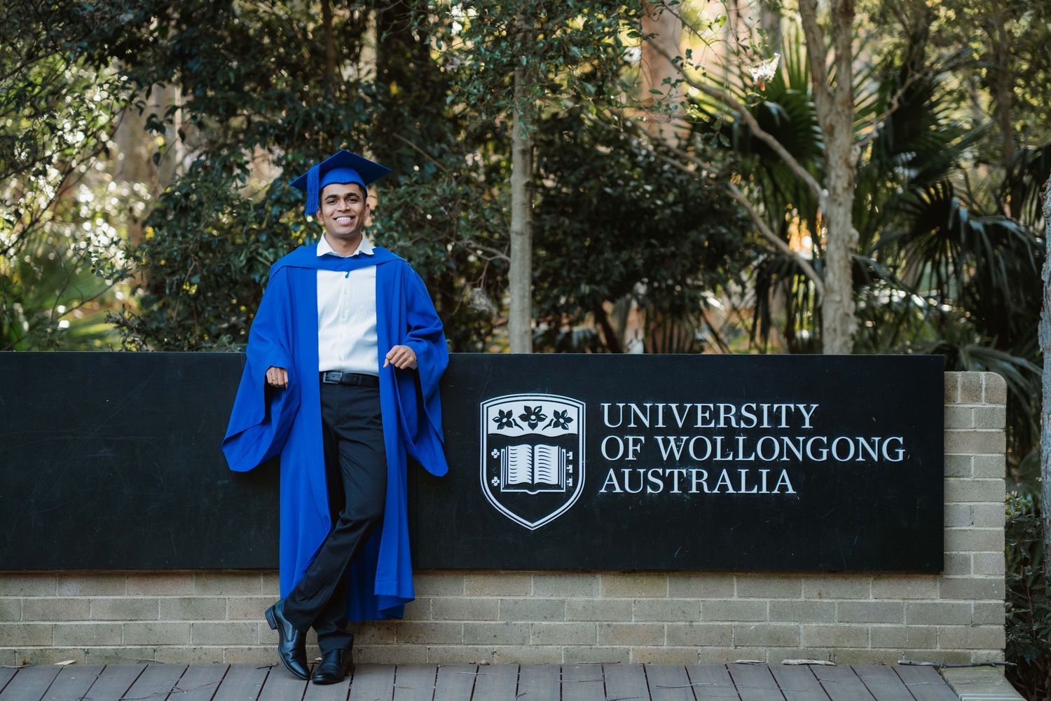 Graduate in a blue gown and cap stands smiling beside the University of Wollongong Australia sign surrounded by trees on graduation day.