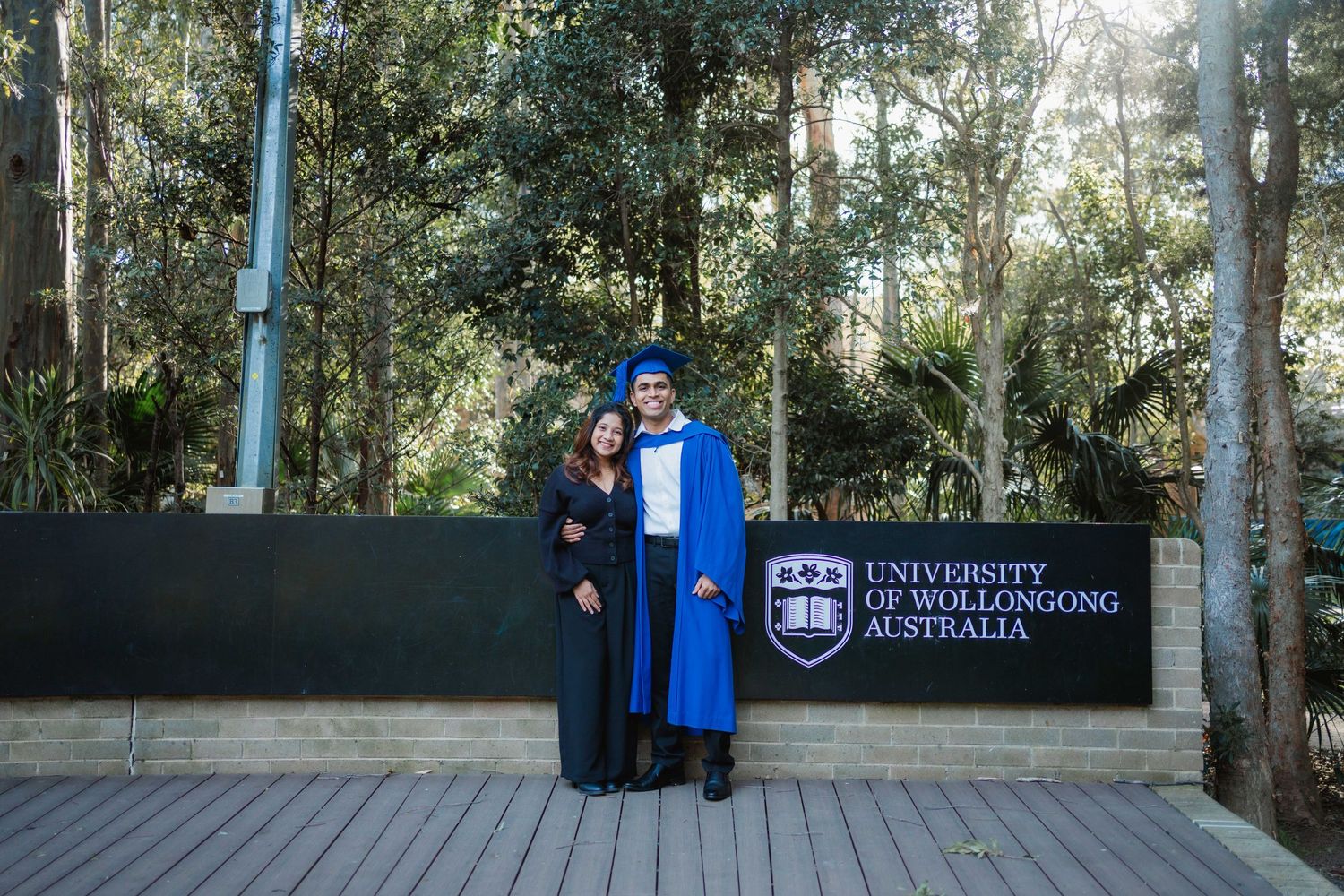 Two University of Wollongong graduates in blue and black gowns pose beside the university sign on a wooden deck surrounded by trees.