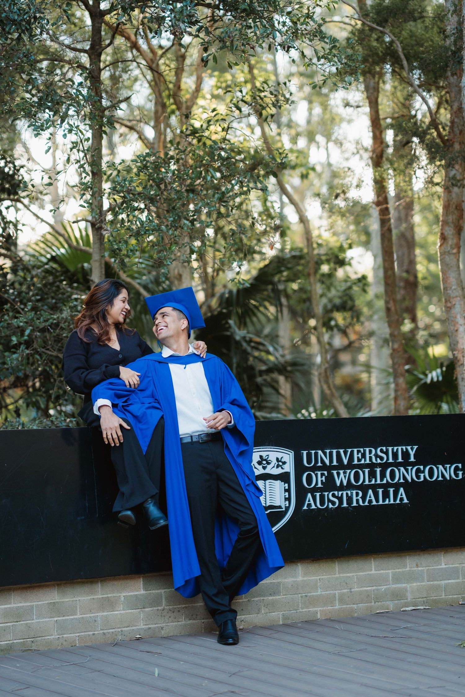 A graduate in a blue gown and cap leans on a wall beside a smiling woman at the University of Wollongong Australia sign.