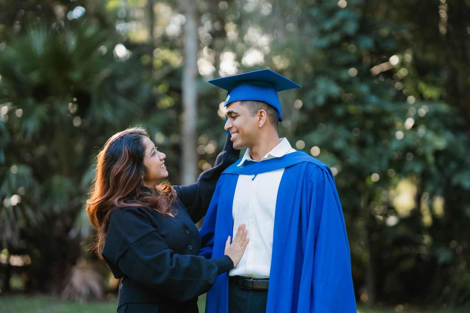 Graduate in a blue gown and cap smiles at a woman in black who holds him warmly while they stand among green trees outdoors.