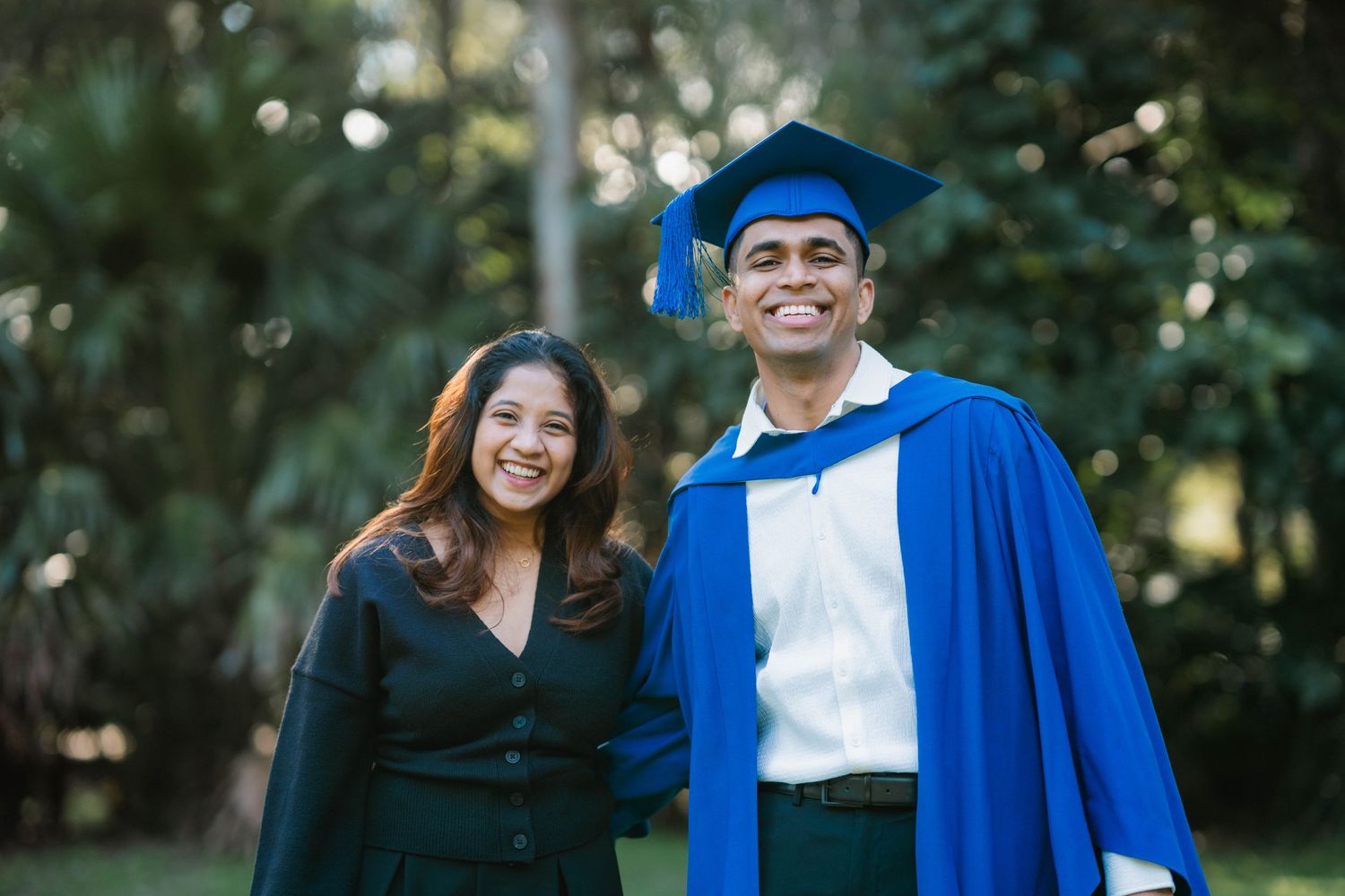 Smiling graduate in a blue gown and cap poses outdoors beside a woman in a black outfit, surrounded by green trees.
