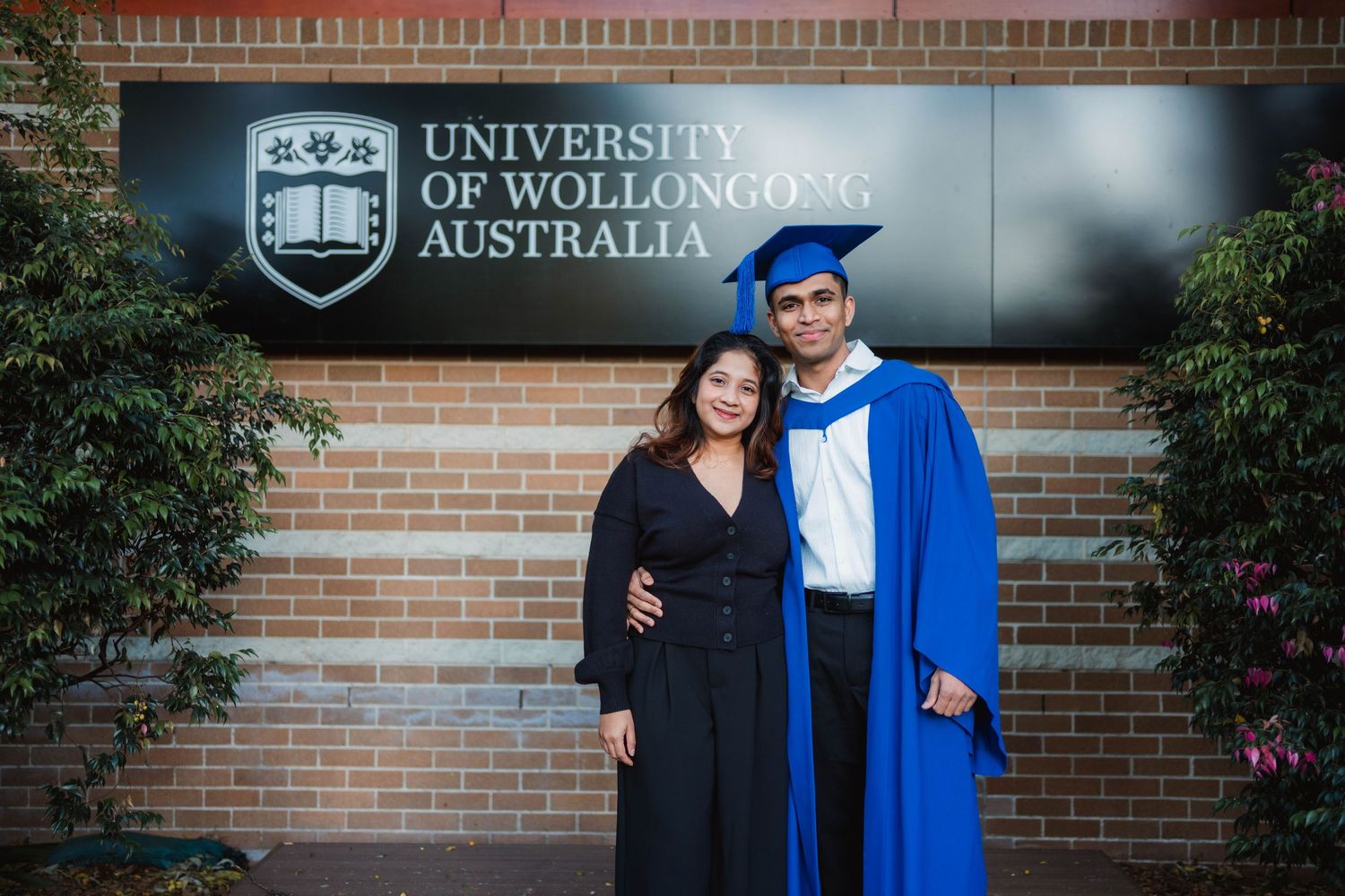 Smiling graduate in a blue gown and cap poses beside a woman in black in front of the University of Wollongong Australia sign.