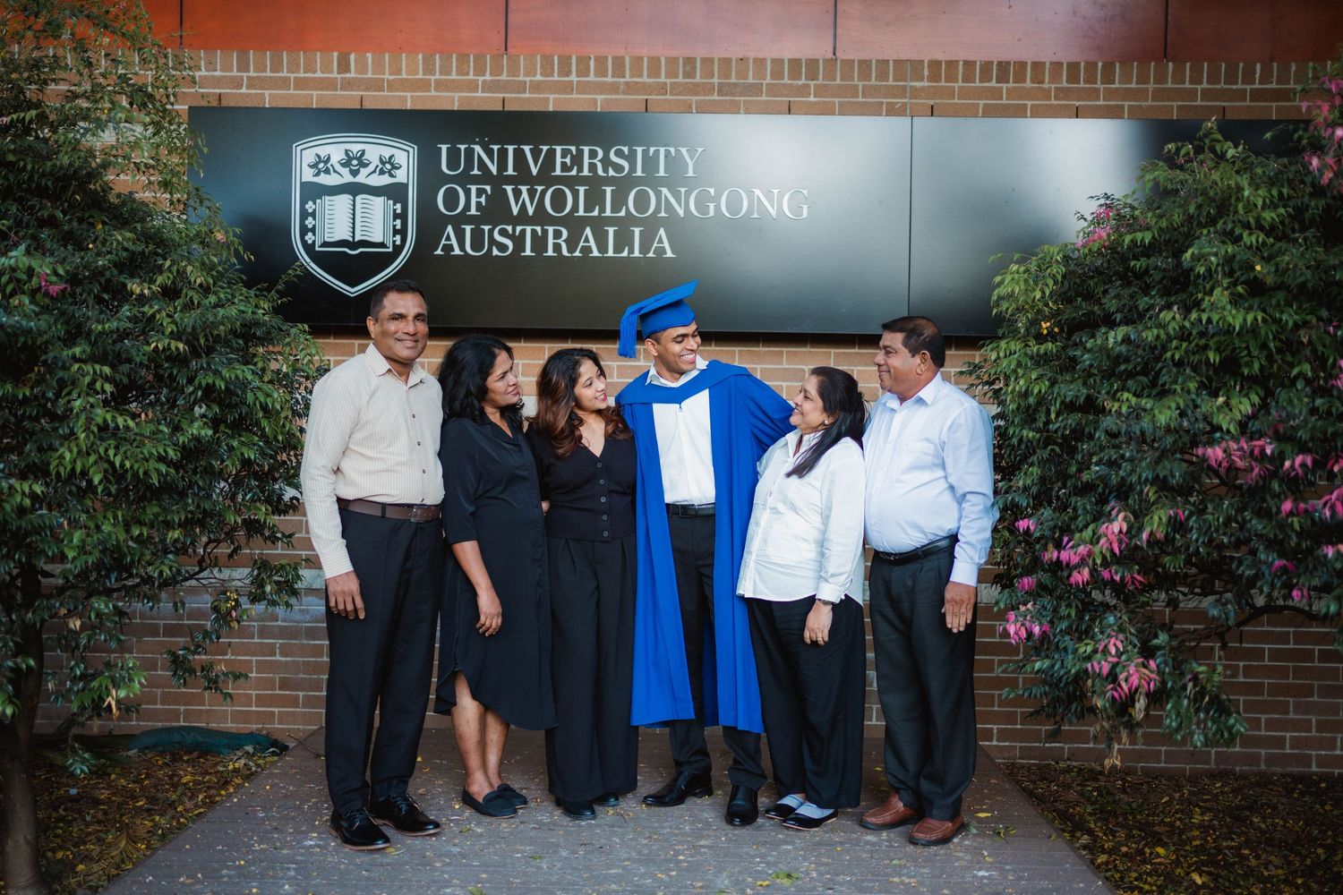 Graduate in blue gown and cap poses with family in front of the University of Wollongong Australia sign beside blooming pink flowers.