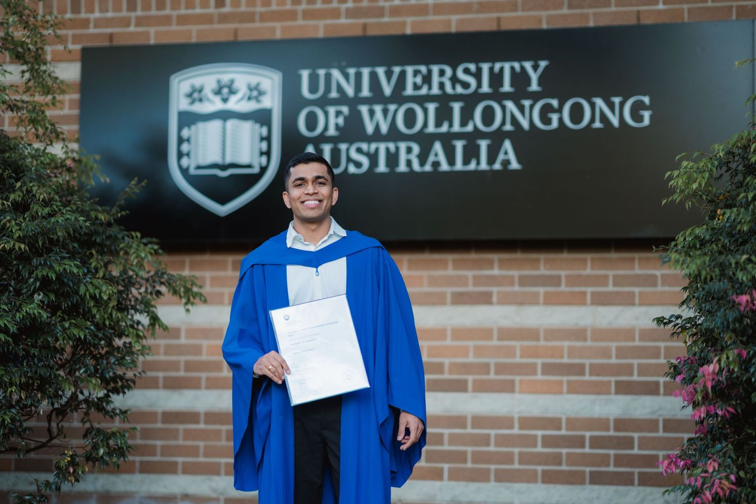 Smiling graduate in a blue gown holds a diploma in front of the University of Wollongong Australia sign on a brick wall.