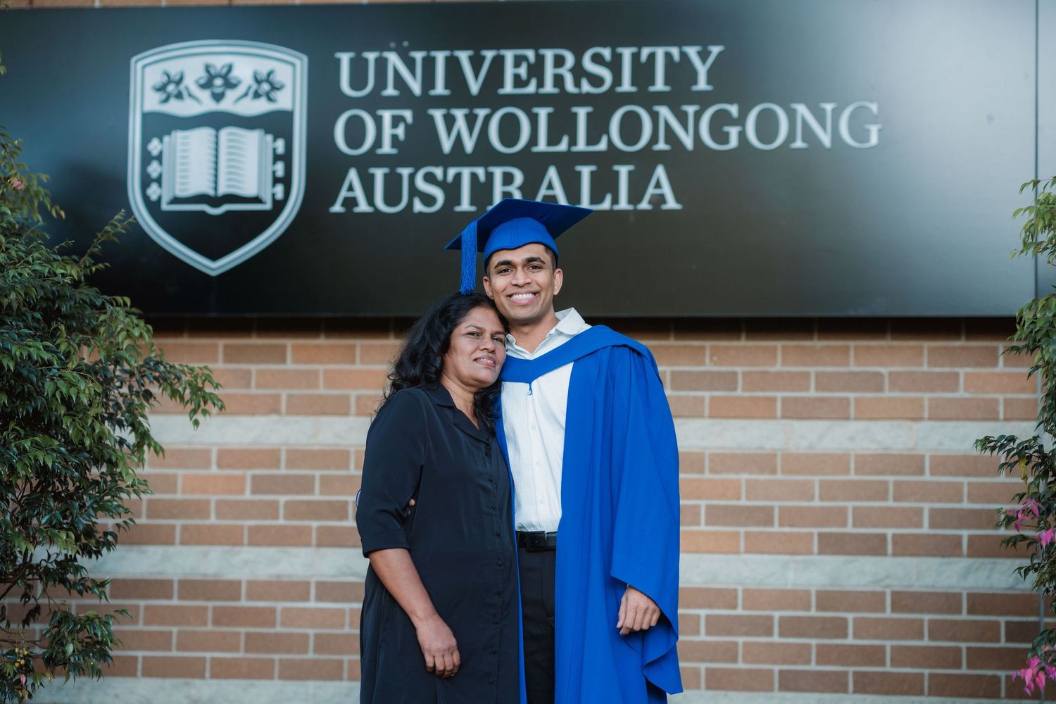 Smiling graduate in blue gown and cap poses with a woman in black in front of the University of Wollongong Australia sign.