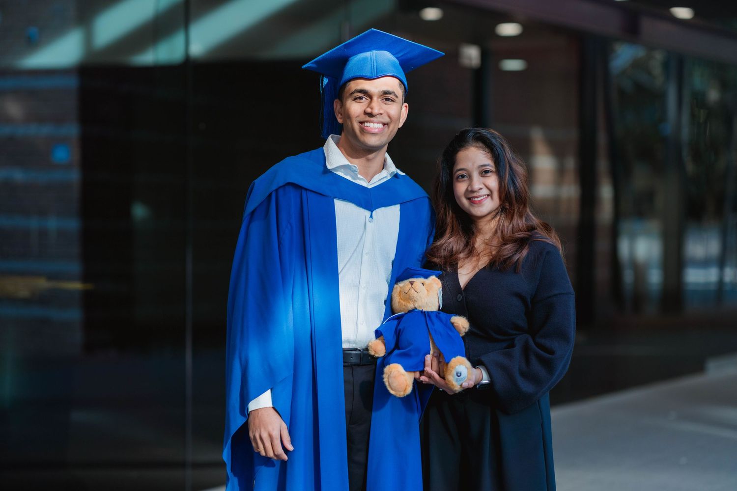Smiling graduate in a blue cap and gown stands beside a woman in a black dress holding a teddy bear in a matching graduation gown.