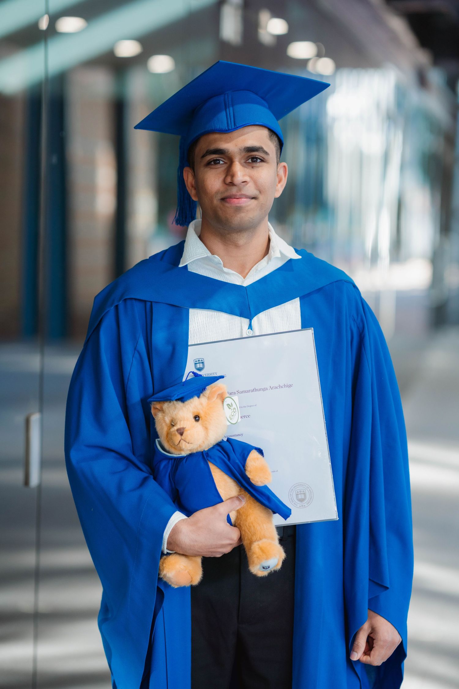Graduate in a blue gown and mortarboard holds a diploma and a teddy bear in matching graduation attire inside a modern university building.