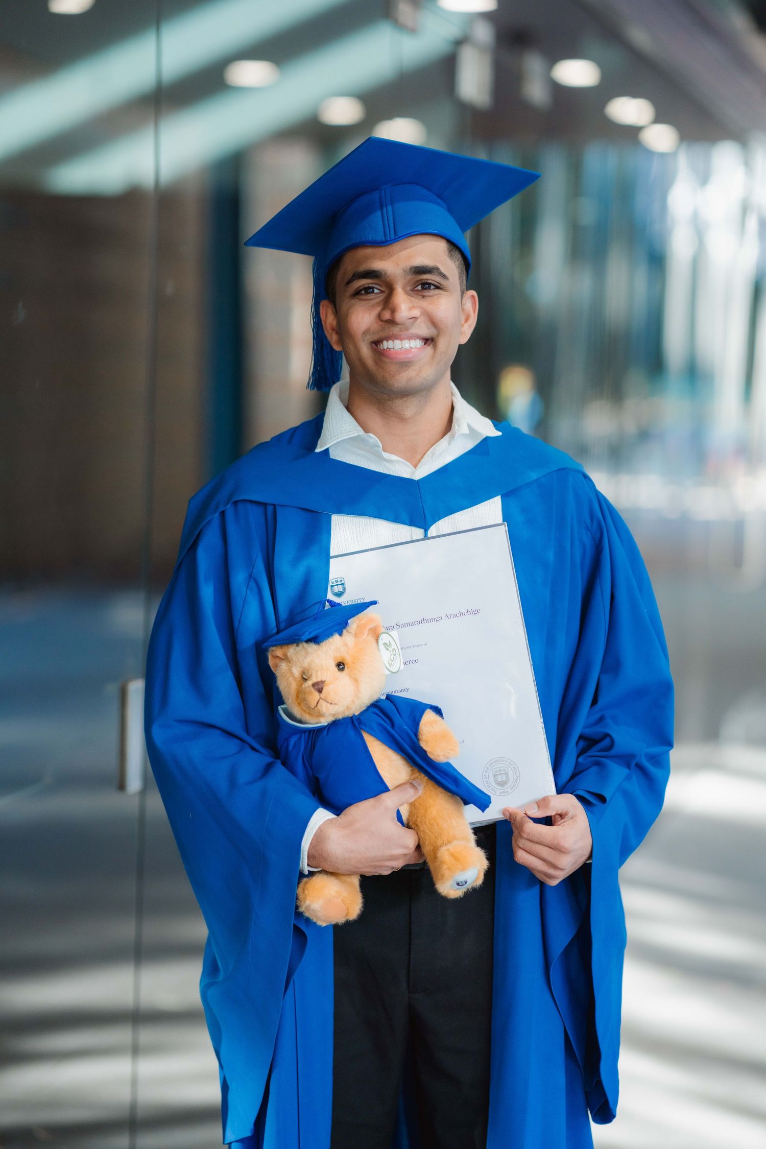 Smiling graduate in a blue cap and gown holds a diploma folder and a teddy bear dressed in matching graduation attire.