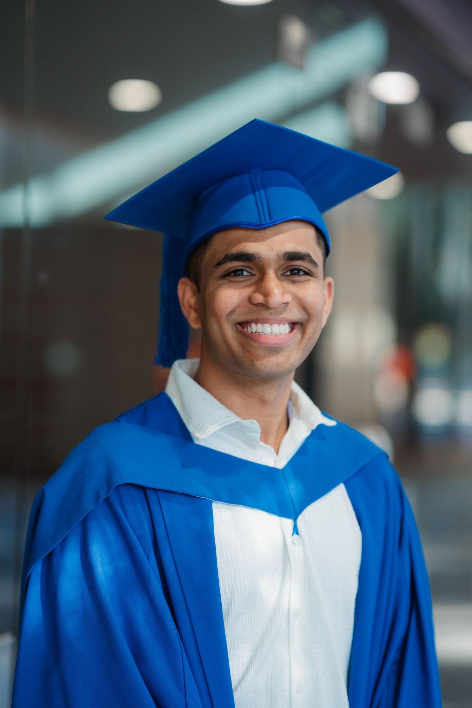 Smiling graduate in a blue graduation cap and gown poses indoors in a bright, modern building with blurred lights in the background.