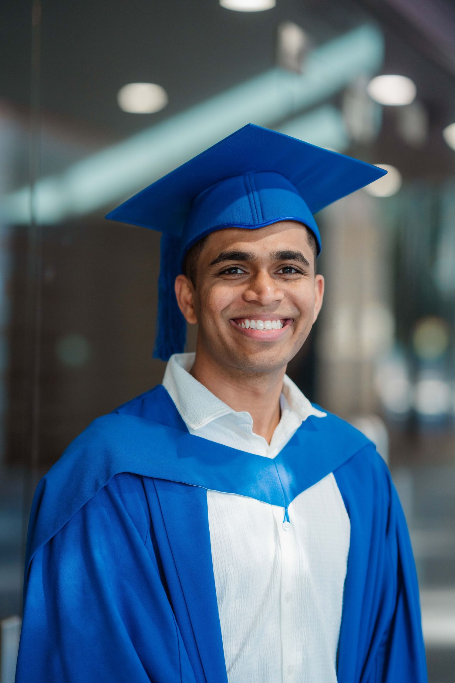 Smiling graduate in a blue cap and gown over a white collared shirt celebrates graduation day in a bright, modern building.