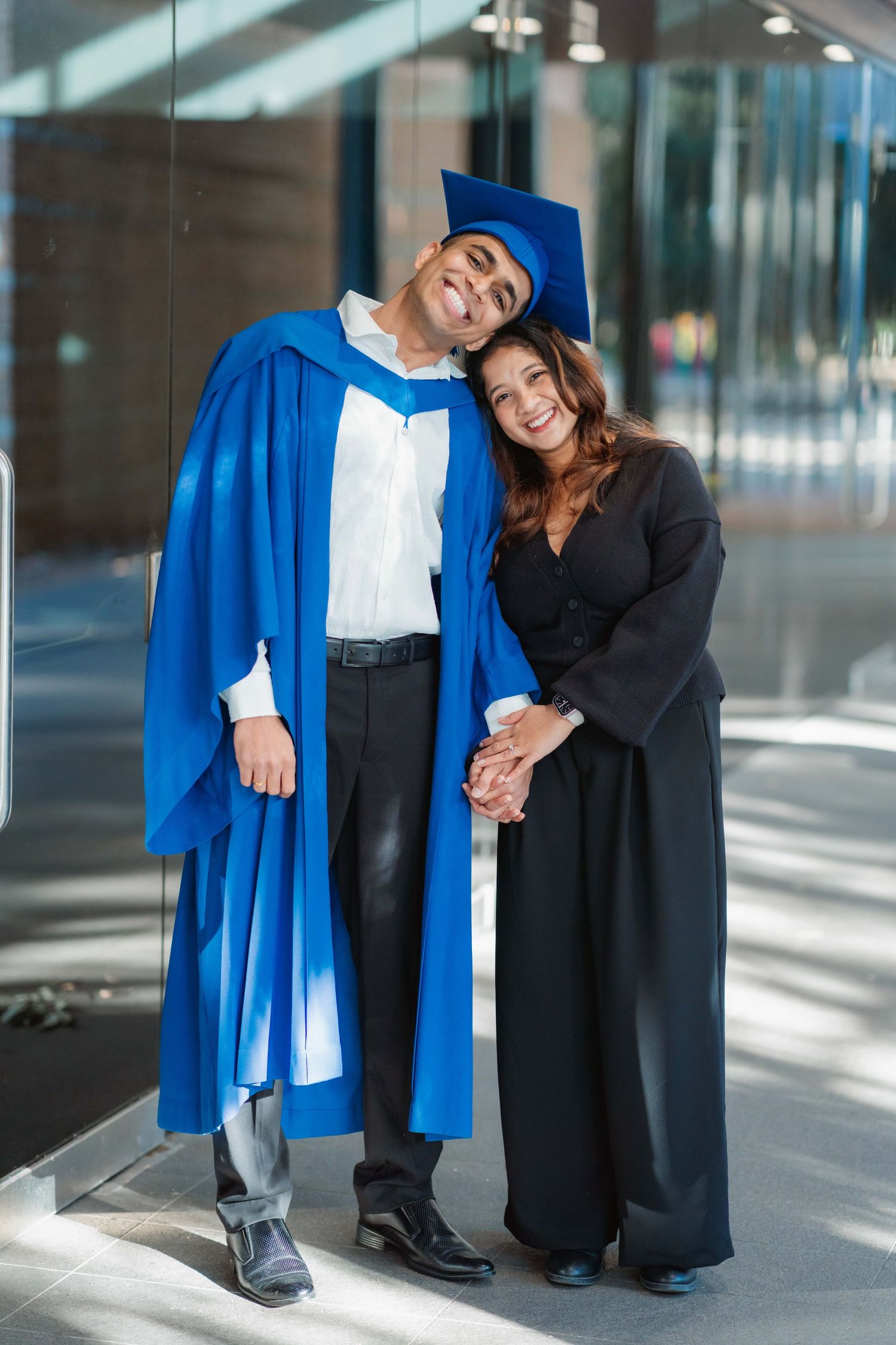 Smiling graduate in a blue gown and cap leans his head on a woman in black as they hold hands in a modern glass building.