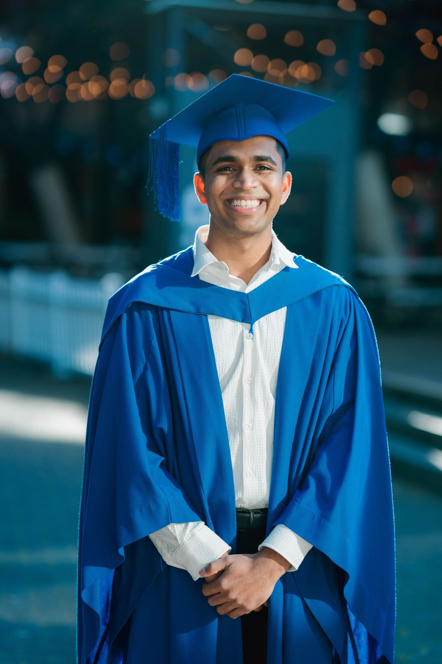 Smiling graduate in a blue gown and matching cap stands with hands clasped, posing outdoors with softly blurred warm string lights behind.