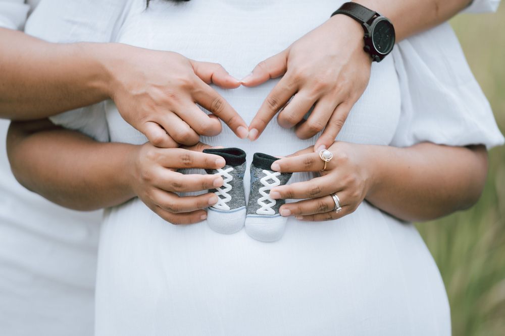 Expecting couple in white outfits forming a heart with their hands over the baby bump while holding tiny gray baby sneakers.