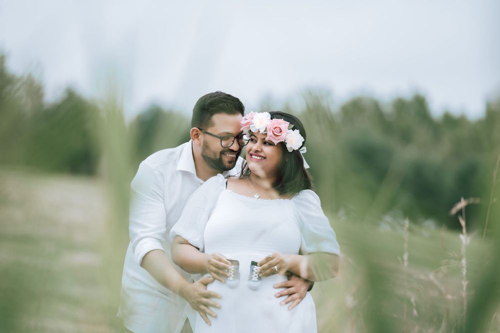 Smiling couple embracing in a grassy field, the woman wearing a white dress and pink floral crown as the man hugs her from behind.