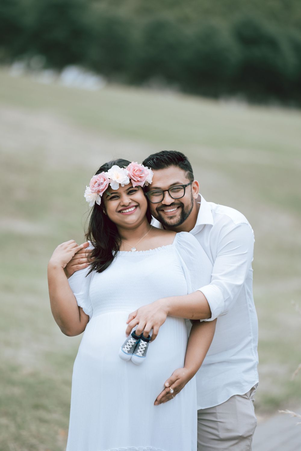 Smiling expectant couple in white outfits pose outdoors on a grassy hillside, holding tiny baby shoes over the mother-to-be's belly.
