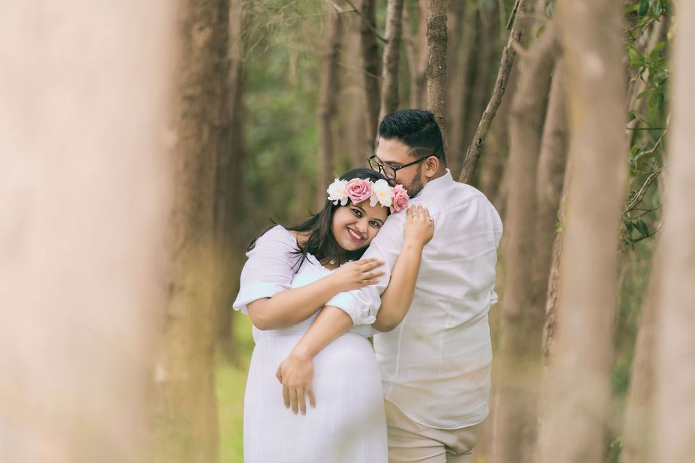 Smiling pregnant woman in a white dress and floral crown embraces her glasses-wearing partner among tall trees in a sunlit forest.