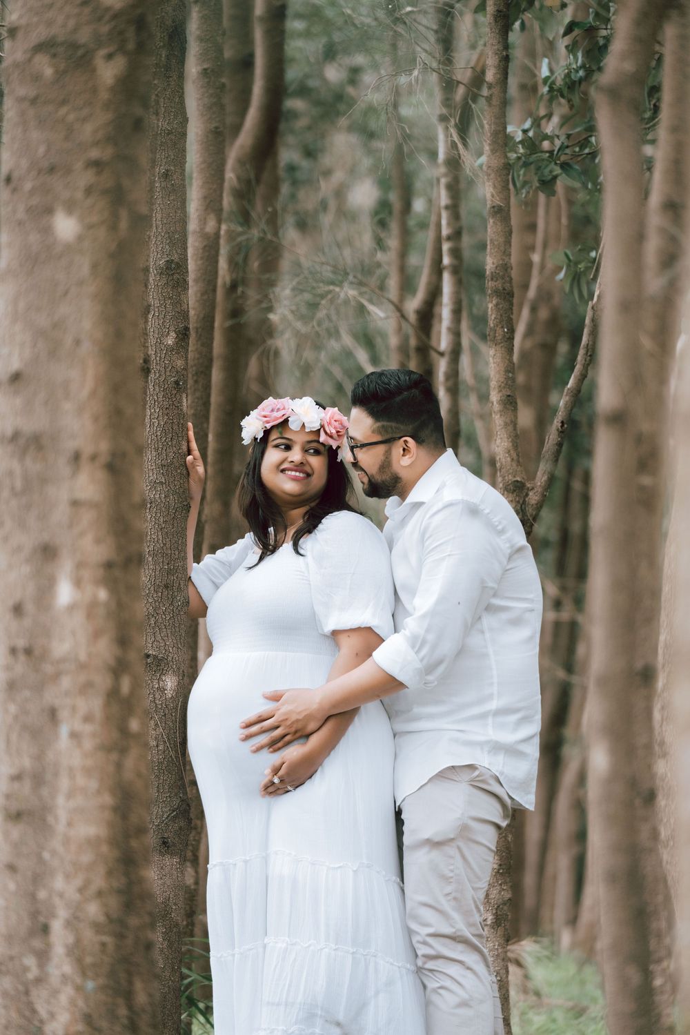 Expecting couple in white outfits embracing in a tree-lined forest, the mother wearing a pink floral crown during a maternity photoshoot.
