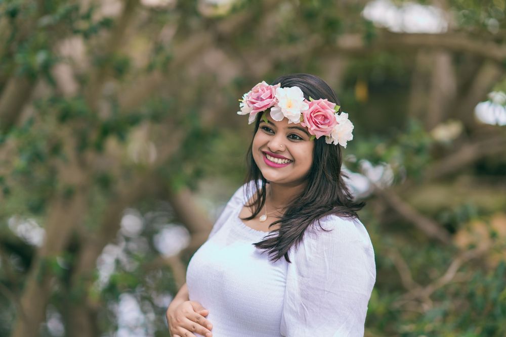 Smiling woman with long dark hair wearing a pink and white floral crown and a white top, standing outdoors among blurred green trees.