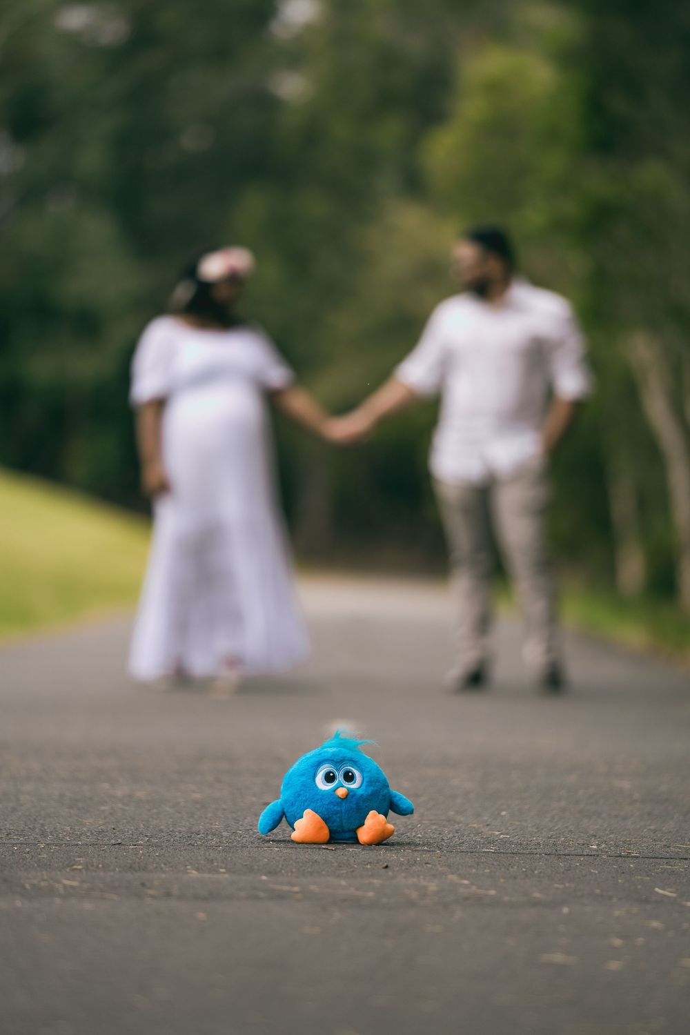 Expecting couple in white outfits holding hands on a park path, with a blue bird plush toy in sharp focus in the foreground.