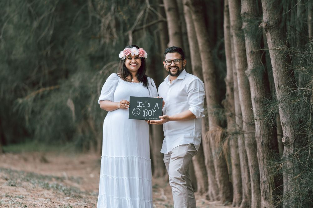 Expecting couple in a pine forest holds a chalkboard sign reading It's a boy, the smiling woman wearing a white dress and floral crown.