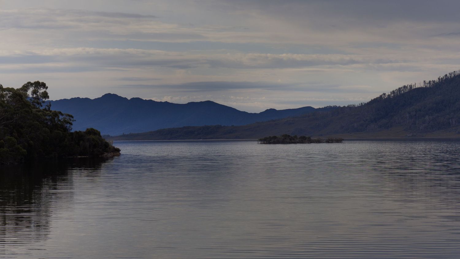 Calm mountain lake reflecting rugged peaks and grassy slopes under a soft, cloudy sky in a remote wilderness landscape.