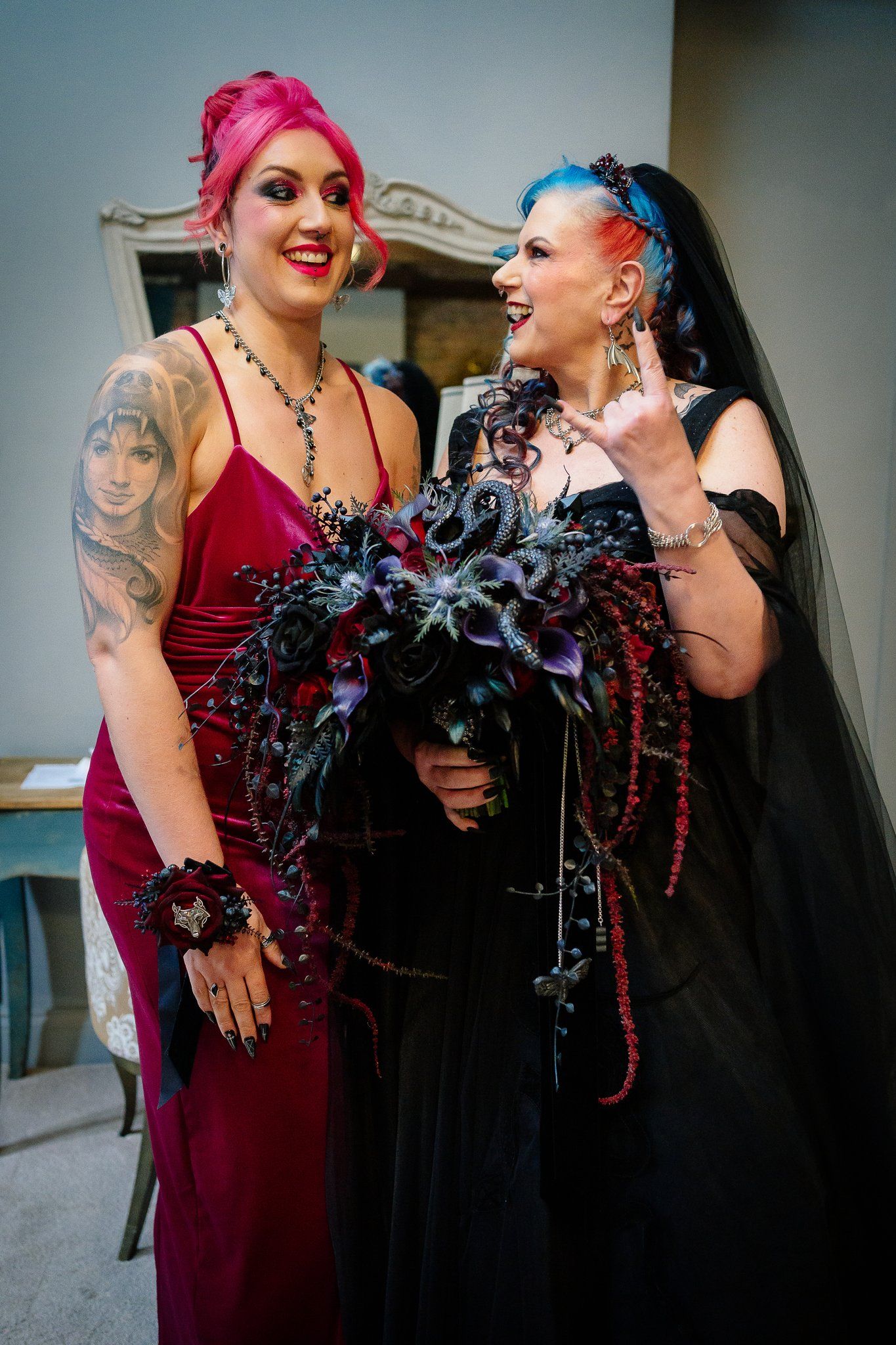 Gothic bride in a black veil laughing with her bridesmaid in a red velvet dress before the ceremony at Le Petit Chateau