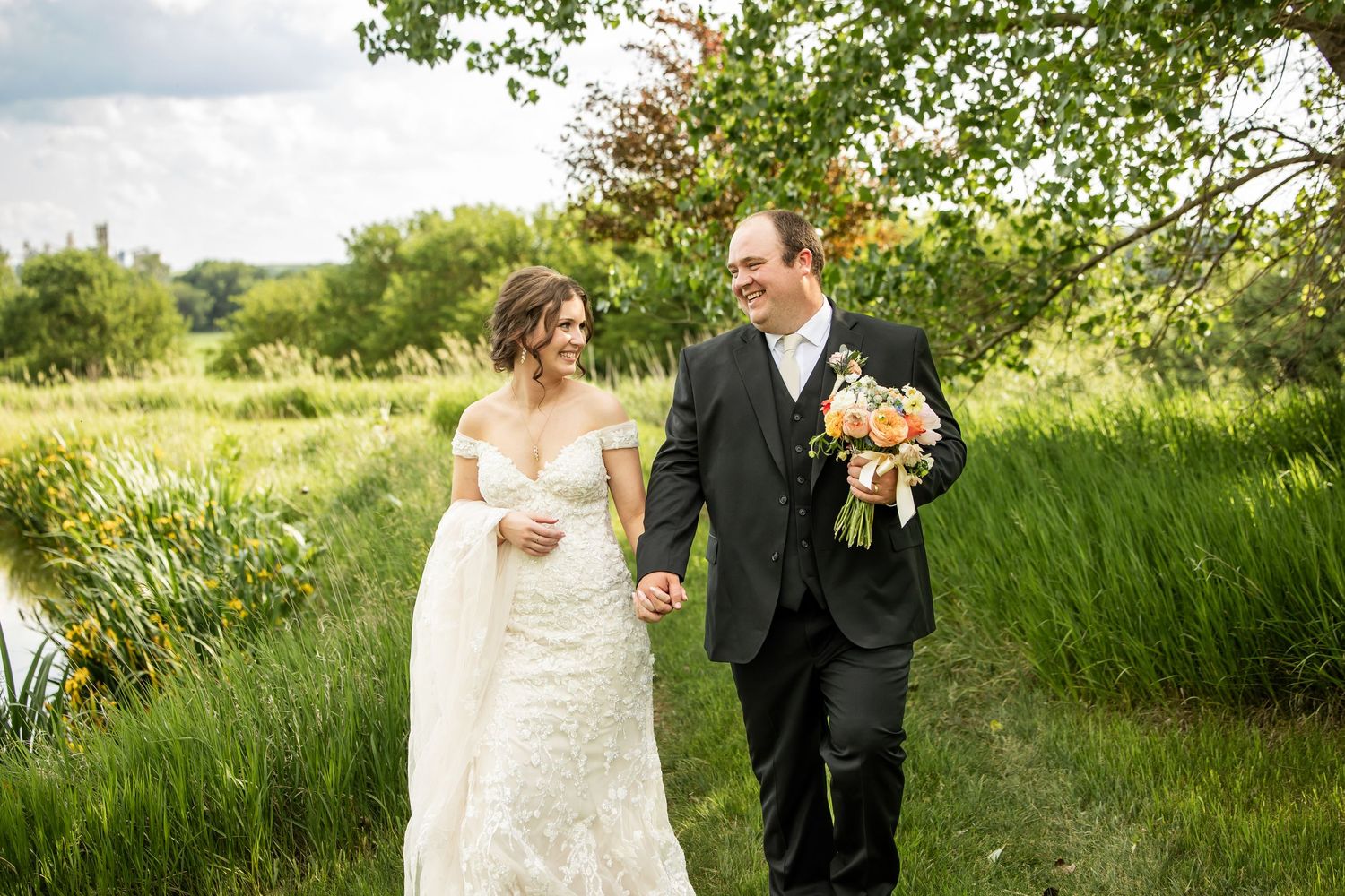 Bride in a lace wedding gown and groom in a dark suit holding hands and walking through a green meadow with a pink bouquet.